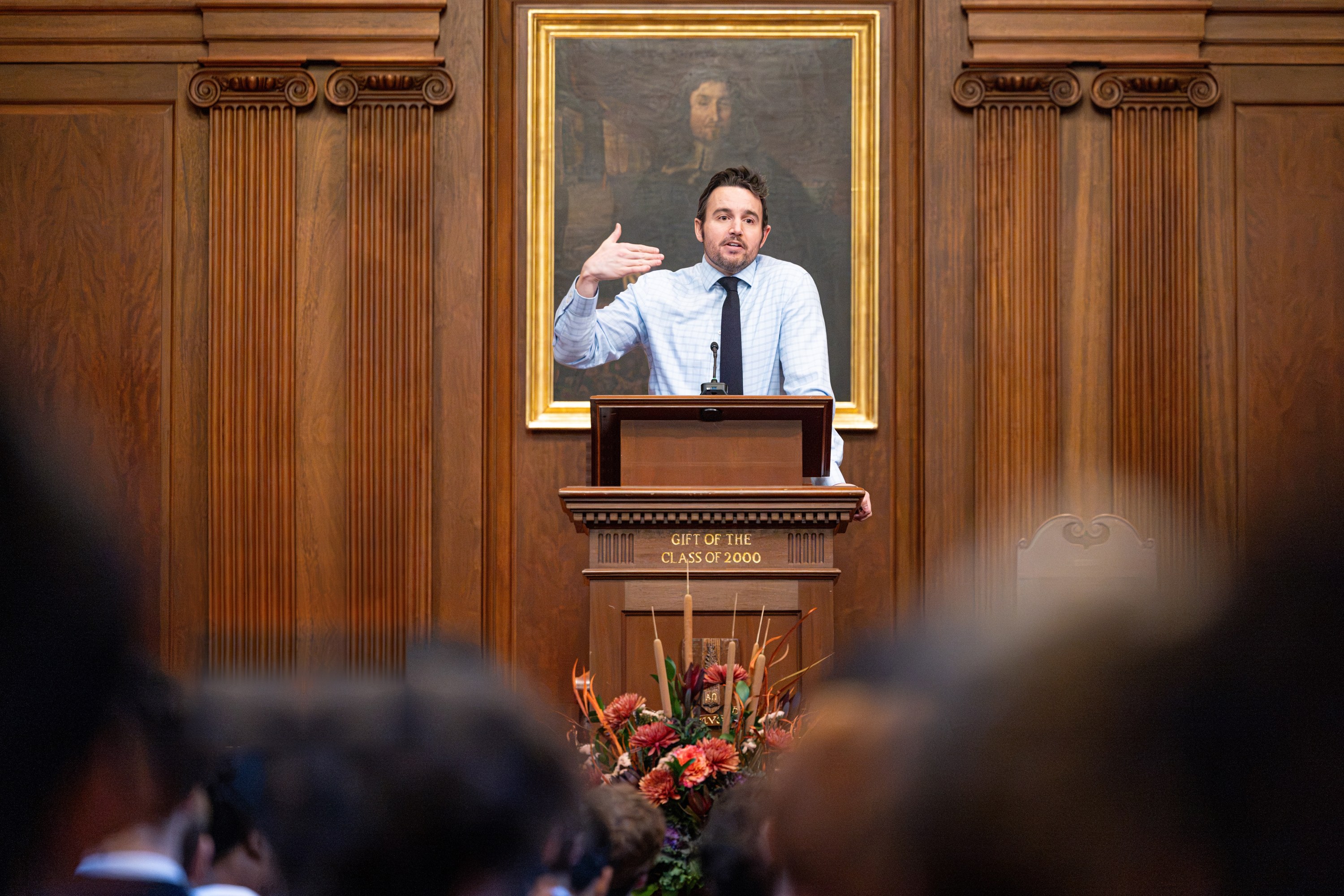 Peter Krause speaking at a wooden lectern, with a framed portrait on the wall behind him and audience members in the foreground.